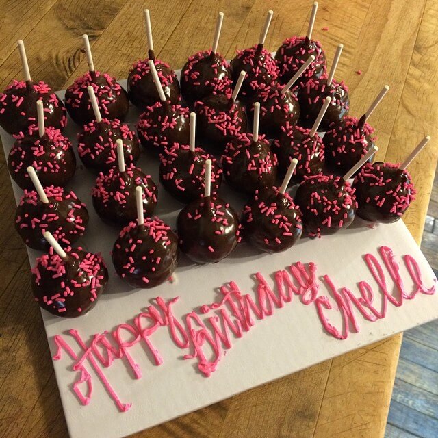 A group of chocolate cake pops with pink sprinkles is arranged neatly on a white board. Decorated with pink icing spelling "Happy Birthday Chelle" on the right side, the festive display looks like a delightful treat from a cozy bakery. The board is set on a wooden surface, adding rustic charm.