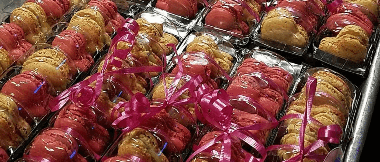Assortment of colorful macarons, some red and some yellow, individually packed in clear plastic containers. The containers, perfect for any bakery or cafe display, are arranged neatly in rows and topped with curly pink ribbons, ready for gifting or sale.