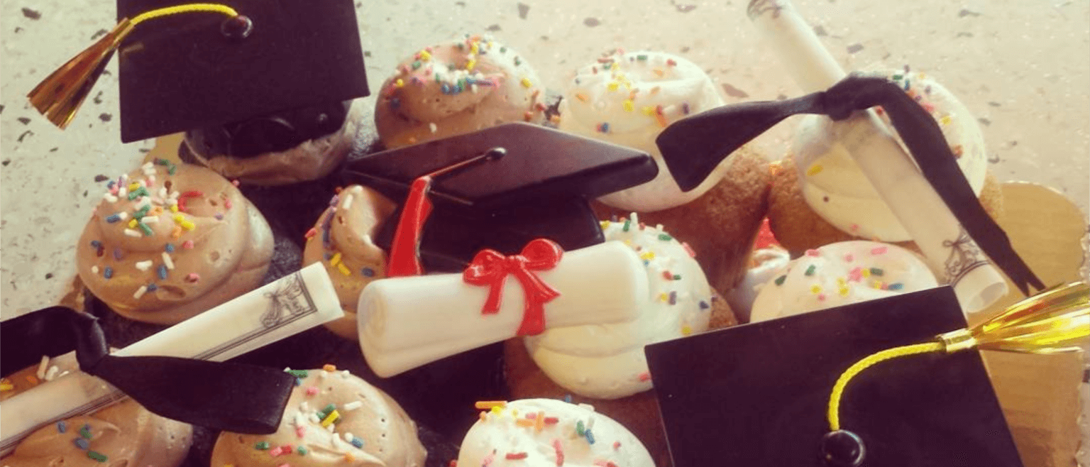 A close-up of celebratory cupcakes from a quaint bakery, topped with chocolate and vanilla frosting, decorated with mini graduation caps and scrolls tied with red ribbons. The cupcakes are sprinkled with colorful confetti.