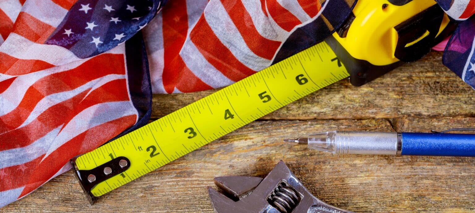 A yellow tape measure, a ballpoint pen, a wrench, and an American flag scarf are laid out on a wooden surface. The tape measure extends across the image, and the colorful flag adds a touch of patriotism to the scene.