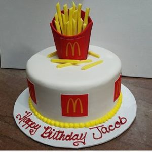 A round white birthday cake, expertly crafted by a bakery, features several McDonald's logos around the sides. On top of the cake is a replica of McDonald's fries in a red container. "Happy Birthday Jacob" is written in red icing at the cake's base.