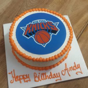 A round cake with white icing and an orange border features the New York Knicks logo in the center. The cake, from a local bakery, is placed on a white square base with orange text that reads, "Happy Birthday Andy." The cake sits proudly on a wooden surface.