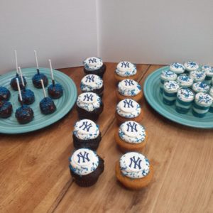 A display of New York Yankees-themed treats on a wooden table in the bakery. There are chocolate cake pops with blue sprinkles, cupcakes with Yankees logo icing, and small dessert cups topped with blue and white decorations. All items are arranged on turquoise plates, like something out of a charming cafe.