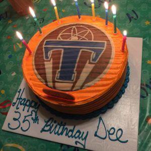 A round birthday cake with orange frosting and a basketball-themed logo on top, crafted by our bakery. The lit candles illuminate the inscription on the white base that reads, "Happy 35th Birthday Dee." The table at our cafe is covered with a green tablecloth adorned with colorful swirls.