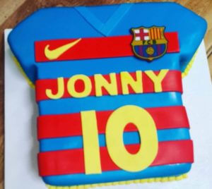 A cake shaped like a blue and red striped FC Barcelona soccer jersey with yellow accents. The jersey, available at our bakery, has the name "JONNY" and the number "10" written in yellow icing. The Nike logo and FC Barcelona crest are also visible on the shirt.