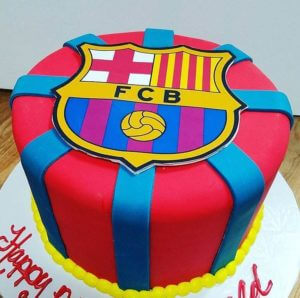 A round cake from our bakery is adorned with the FC Barcelona logo on top. Primarily red with blue stripes running vertically and horizontally, it rests on a white plate bordered by yellow frosting. Partial text written in red is visible on the plate's edge, making it perfect for any cafe display.