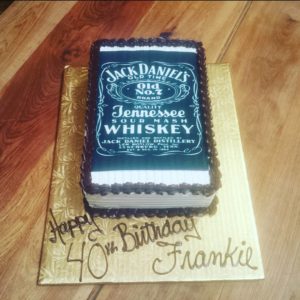 A bakery masterpiece, this birthday cake designed to look like a Jack Daniel's whiskey label sits elegantly on a wooden surface. "Happy 40th Birthday Frankie" is written in chocolate on the golden cake board. The cake from our cafe has a white base with black icing around the edges.