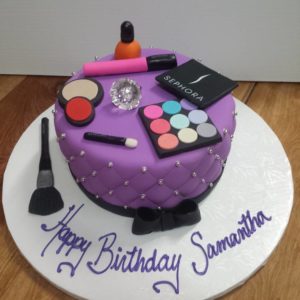 A round, purple birthday cake from our charming bakery is decorated with makeup-themed fondant toppers including a brush, eyeshadow palette, lipstick, and nail polish bottle. Adorned with small silver beads and icing that reads "Happy Birthday Samantha," this cake is perfect for any celebration.