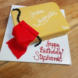 A birthday cake designed to look like a beige Christian Louboutin shoebox with black rope handles and a red cloth. Crafted by our cafe bakery, it includes a handwritten birthday message in red icing: "Happy Birthday Stephanie!" The cake is placed on a white textured board on a wooden surface.