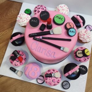 A decorated birthday cake and cupcakes themed around makeup, crafted by our bakery. The main cake is pink with brushes, lipsticks, and eyeshadow on top, and "Clarisa" in the center. Surrounding cupcakes are similarly adorned; one reads "18". Visit our cafe for delicious treats with a creative twist.