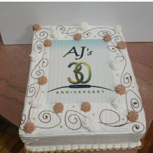 A rectangular white cake with decorative brown and white frosting swirls and dollops, crafted by a local bakery, features a central image with the text "AJ's 30th Anniversary 1988-2018." The image includes a large number "30" and is framed by small gold beads.