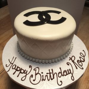 A white birthday cake with the Chanel logo in black on top, crafted by our boutique bakery. The cake features a quilted pattern and a border of white pearl-like beads. "Happy Birthday Rose" is elegantly written in chocolate on the white cake board. Perfect for any cafe celebration!