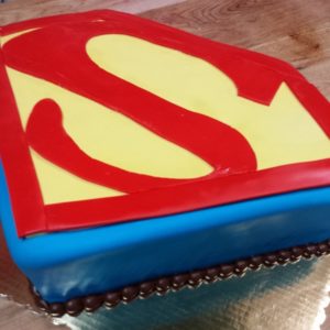 A cake from our bakery, decorated with a blue base and topped with a red and yellow Superman logo, featuring a red 'S' on a yellow background. Sitting proudly on a wooden surface in our cozy cafe, the cake is trimmed with chocolate balls around the bottom edge.