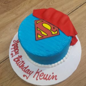 A birthday cake decorated with a Superman theme sits proudly in the bakery. The top is blue with a red and yellow Superman logo, and a piece of red fondant resembling a cape. The cake sits on a white base with "Happy Birthday Kevin" written in red icing along the edge.