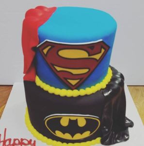 Two-tiered fondant cake from our bakery themed after superheroes: the top tier features a blue base with the Superman logo and a red cape, while the bottom tier has a black base with the Batman logo and a black cape. Yellow fondant bands separate the tiers. Perfect for any superhero-themed party at our cafe!
