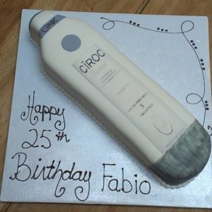 A rectangular birthday cake designed to look like a Cîroc vodka bottle, with white icing and gray accents, sits on display at the bakery cafe. The message "Happy 25th Birthday Fabio" is written in cursive on the cake board. The background features a wooden surface with decorative flourishes.