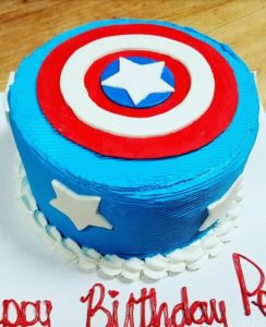 A round, blue birthday cake with a red, white, and blue design resembling Captain America's shield on the top sits proudly at our bakery. The sides are adorned with white star-shaped fondant pieces, and the base has a fluffy white border. "Happy Birthday" is elegantly written in red icing.