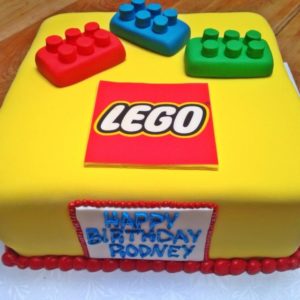 A square, yellow birthday cake from our bakery decorated to resemble LEGO bricks. The top features a red, blue, and green brick along with the LEGO logo. The front of the cake has a white panel bordered in red icing with the text "HAPPY BIRTHDAY RODNEY" written in blue icing.