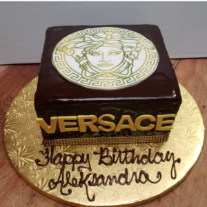 A square cake with a glossy chocolate finish sits on a gold platter. The top features a Versace logo with the Medusa head in white and gold. The side of the cake has "VERSACE" written in gold. This bakery masterpiece has "Happy Birthday Aleksandra" written in chocolate script on the platter.