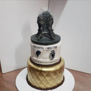 A two-tiered wedding cake crafted by our bakery with a "Game of Thrones" theme. The bottom tier is gold with a quilted pattern, and the top tier is white with the text "House Kuruvilla," "House Ittiyopp," and "Wedding is Coming." The cake from our cafe is topped with an Iron Throne replica.