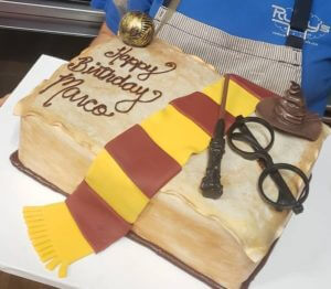 A rectangular cake designed to look like an open book with the message "Happy Birthday Marco." Crafted at our cafe, this bakery masterpiece is decorated with a Gryffindor scarf, a wand, round glasses, the Sorting Hat, and a golden snitch, all from the Harry Potter series.