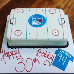 This rectangular birthday cake, crafted by our bakery, is designed to resemble an ice hockey rink. The top features the New York Rangers logo at center ice. It reads "Happy 30th Bobby!" in red icing and includes a blue fondant jersey with "Dr Rico 94" on it. The cake sits on a wood surface.